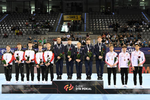 The U.S. team wins gold in the junior men's Team Challenge at the 2023 DTB Pokal, with Germany winning silver (left) and Turkey winning bronze (right). From left to right: Alex Nitache (GymTek), Zach Green (Stanford Boys), Danila Leykin (EVO), Solen Chiodi (Mini-Hops), and Max Odden (North Valley). (© Filippo Tomasi)